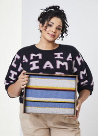 Young woman with curly hair holds a framed textile art piece with blue, yellow, and red stripes. She wears a black "I AM" sweater, exuding confidence.