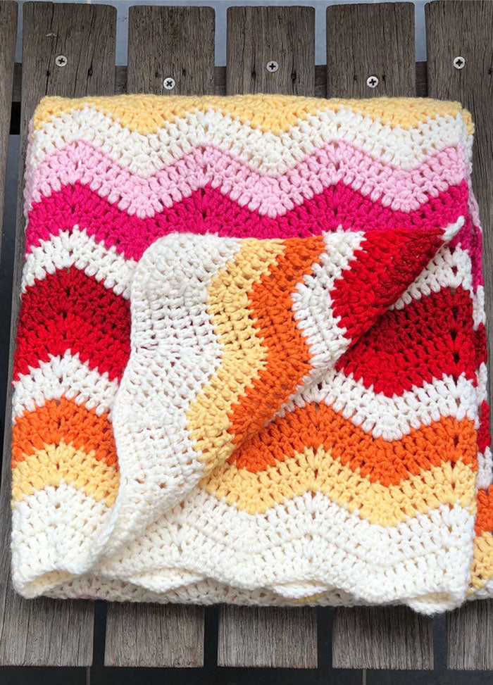 Miami Baby Blanket – folded crocheted baby blanket with a vibrant zigzag stripe pattern in white, yellow, pink, red, and orange hues, displayed on a wooden surface.