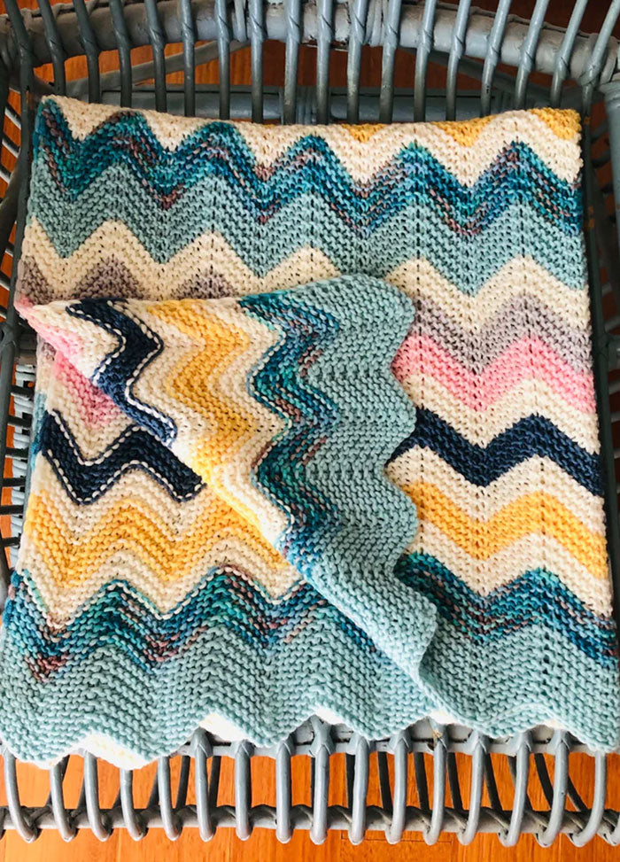 Miami Baby Blanket – multicolored knitted zigzag baby blanket in shades of blue, yellow, pink, and white, casually draped over a wicker chair for a cozy and vibrant nursery touch.