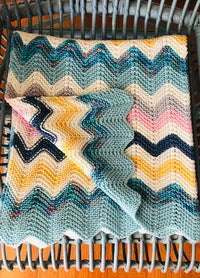 Miami Baby Blanket – multicolored knitted zigzag baby blanket in shades of blue, yellow, pink, and white, casually draped over a wicker chair for a cozy and vibrant nursery touch.