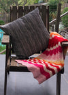 Miami Baby Blanket – vibrant baby blanket with wavy knit patterns in pink, red, orange, and white, draped over a wooden chair with a black-and-white striped cushion, set against a lush poolside backdrop.