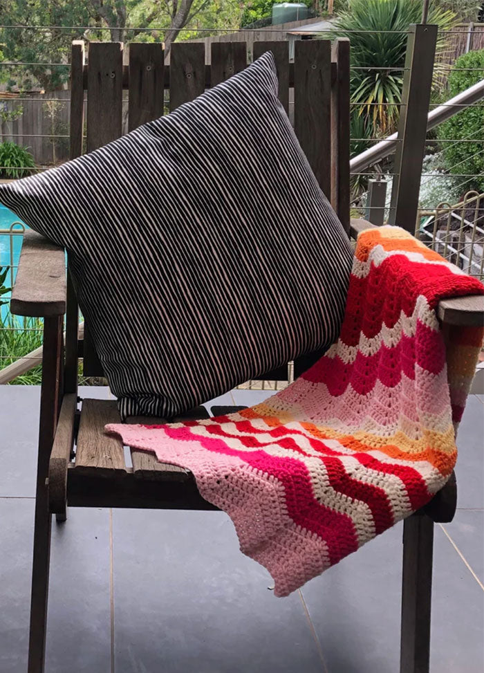 Miami Baby Blanket – vibrant baby blanket with wavy knit patterns in pink, red, orange, and white, draped over a wooden chair with a black-and-white striped cushion, set against a lush poolside backdrop.