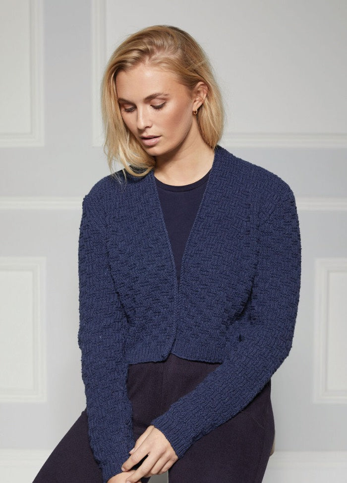 A woman with blonde hair, wearing a textured navy blue cardigan over a matching blue top, stands against a neutral, paneled background. She appears thoughtful.