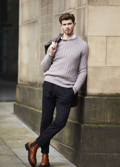 Man leans against a stone wall, wearing a gray sweater, dark pants, and brown boots, exuding a relaxed, confident vibe in an urban setting.