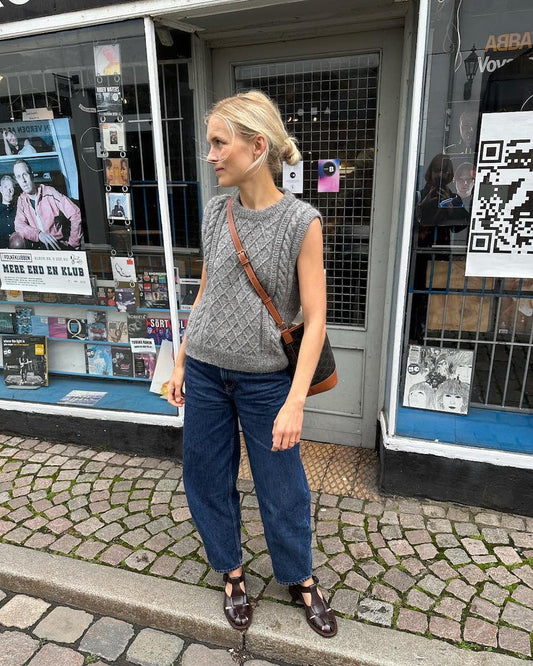 A woman in a grey sleeveless sweater and blue jeans stands on a cobbled street, carrying a brown shoulder bag. She is outside a store featuring various posters.