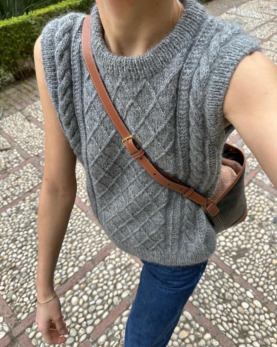 Person in a grey cable-knit sleeveless sweater and blue jeans. They carry a brown crossbody bag while walking on a textured stone path.