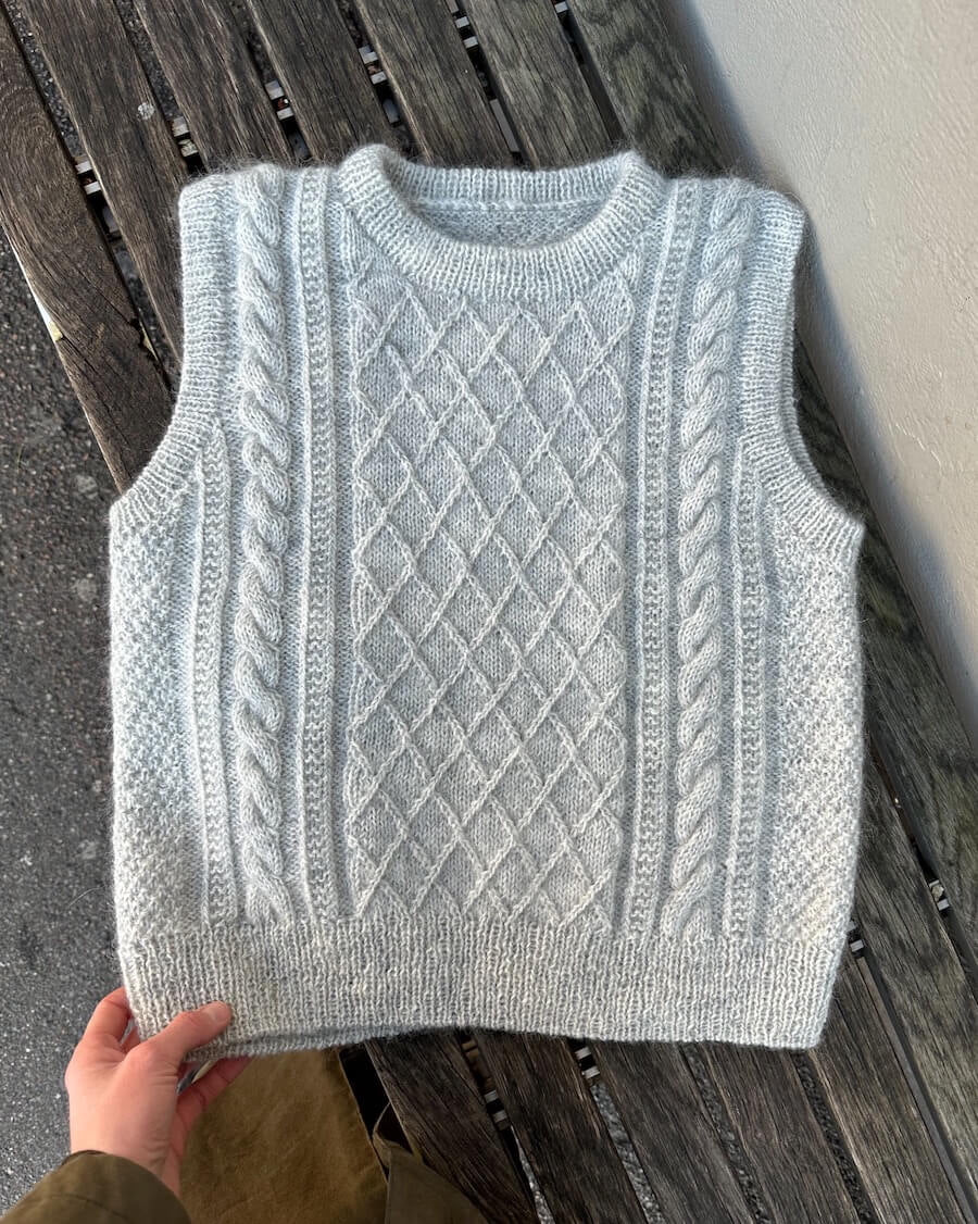 Grey knitted vest with diamond and cable patterns, laid flat on a wooden bench. A hand holds the hem, showcasing the intricate texture and cozy design.