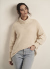 Woman in a cozy cream sweater and light blue jeans leans against a plain wall, exuding a calm and relaxed demeanor with a subtle smile.