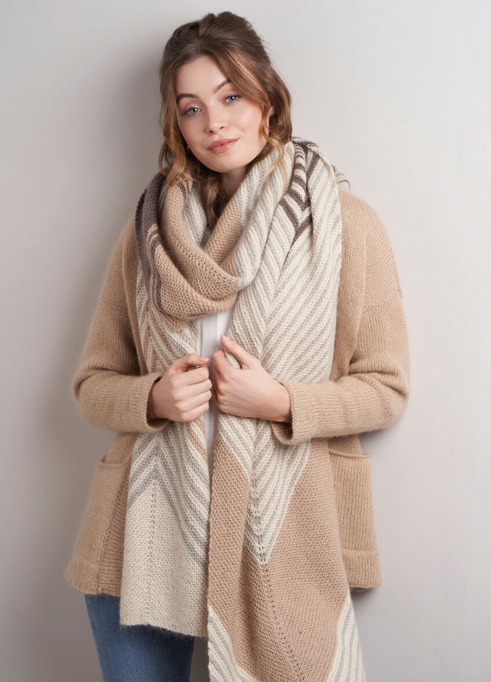 Young woman with light brown hair, wearing a cozy beige sweater and a large striped knitted scarf, stands against a neutral background, exuding warmth.