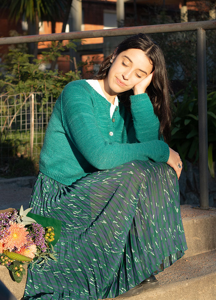Person seated on outdoor steps wearing a teal Montevideo Cardigan and patterned pleated skirt, with flowers beside them.