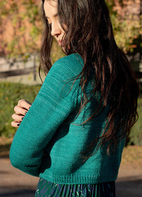 Person with long dark hair wearing a hand-knit teal Montevideo Cardigan with ribbed hem, standing outdoors near greenery.