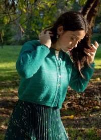 Person wearing green Montevideo Cardigan with front buttons and pleated skirt, standing outdoors among trees and greenery.
