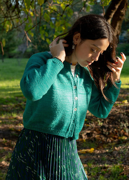 Person wearing green Montevideo Cardigan with front buttons and pleated skirt, standing outdoors among trees and greenery.
