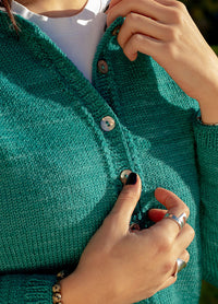 Close-up of person buttoning teal Montevideo Cardigan with black nail polish, rings, and bracelet, against blurred background.
