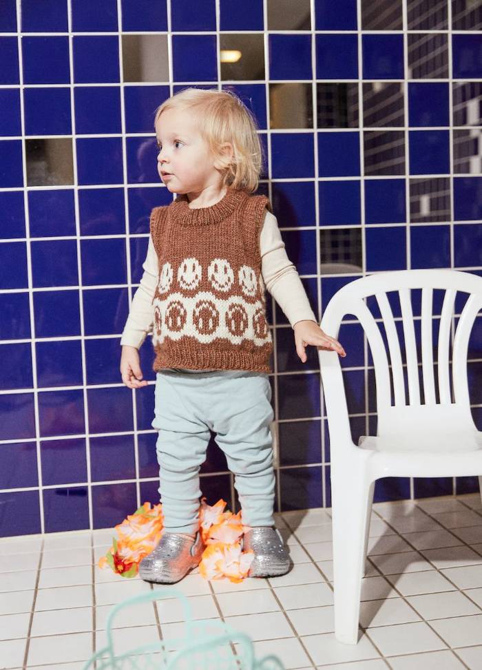 A toddler with blonde hair stands on tiled flooring, wearing a brown knit vest with smiley faces and light blue pants. Nearby is a white plastic chair. The mood is playful.