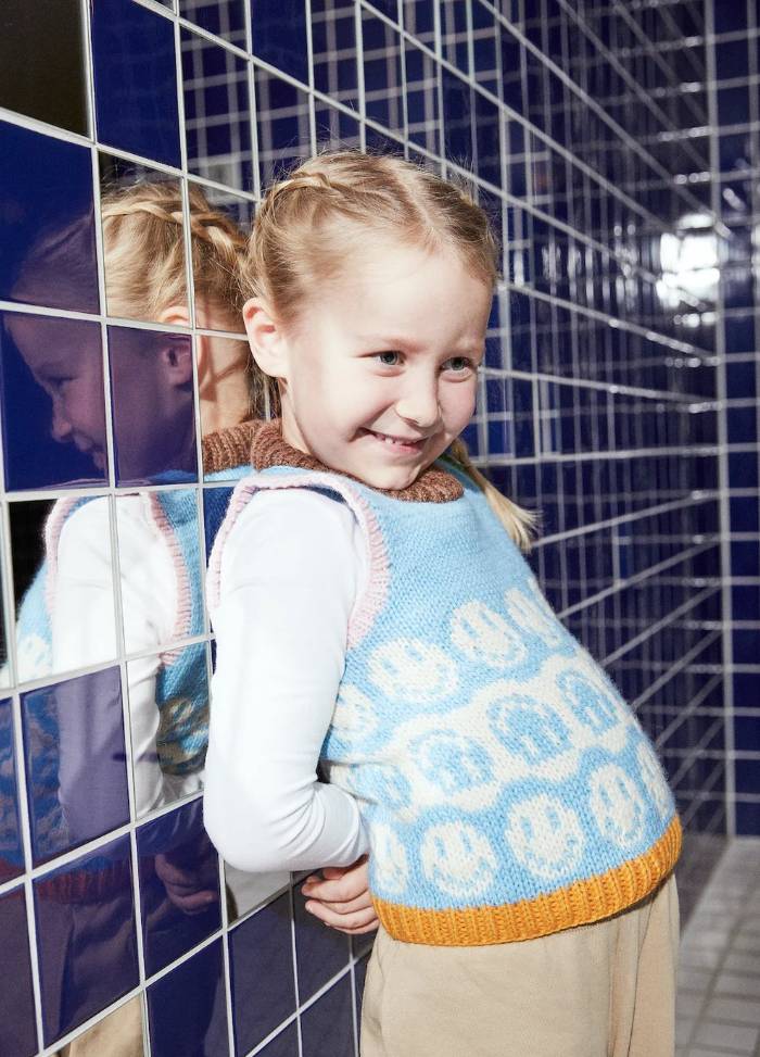A young girl with braided hair smiles while leaning against glossy blue tiles. She's wearing a blue sweater vest with smiley faces, conveying joy and playfulness.