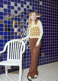A young woman smiles, wearing a yellow sweater vest with smiley faces and brown pants, standing by a white plastic chair against blue tiled walls.