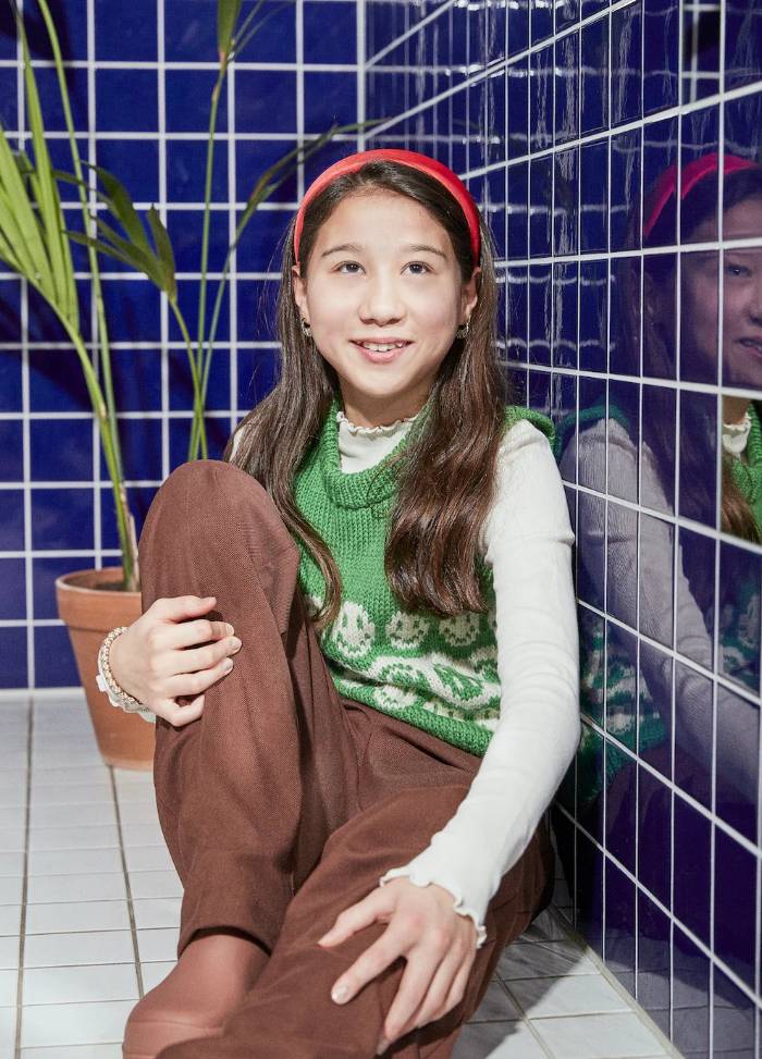 A young girl sits against a blue tiled wall, wearing a green sweater vest, brown pants, and a red headband. She smiles warmly, creating a cheerful atmosphere.