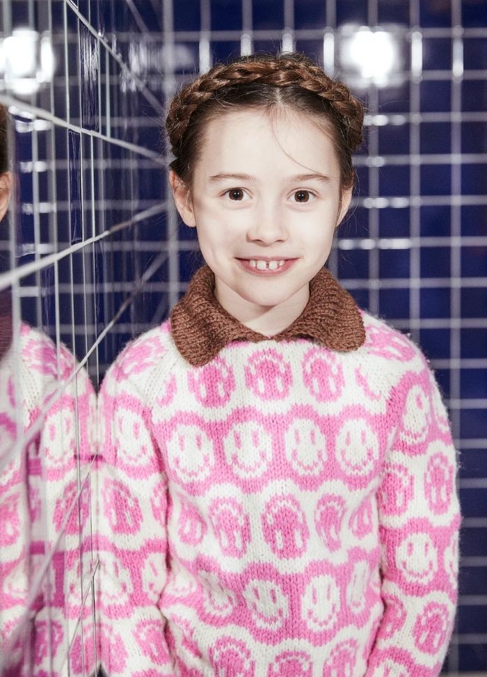 Young girl with braided hair smiles warmly, wearing a pink sweater with smiley faces and brown collar. Reflected in blue tiled wall, evoking cheerful vibe.