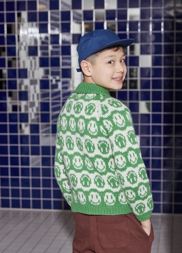 Young boy smiling, wearing a blue cap and a green sweater with white smiley faces, stands against a dark blue tiled wall, creating a playful tone.