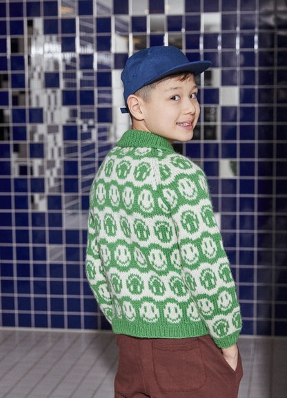 Young boy smiling, wearing a blue cap and a green sweater with white smiley faces, stands against a dark blue tiled wall, creating a playful tone.