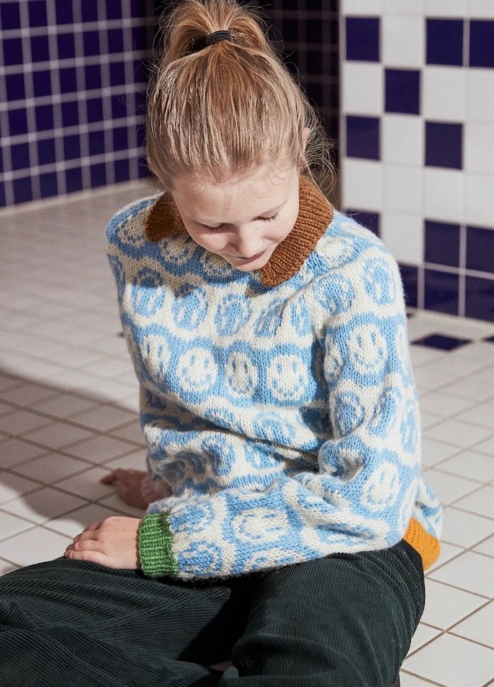 Young person with a ponytail sits on white tiled floor, wearing a blue sweater with smiley patterns and dark pants. Background features blue and white tiles.
