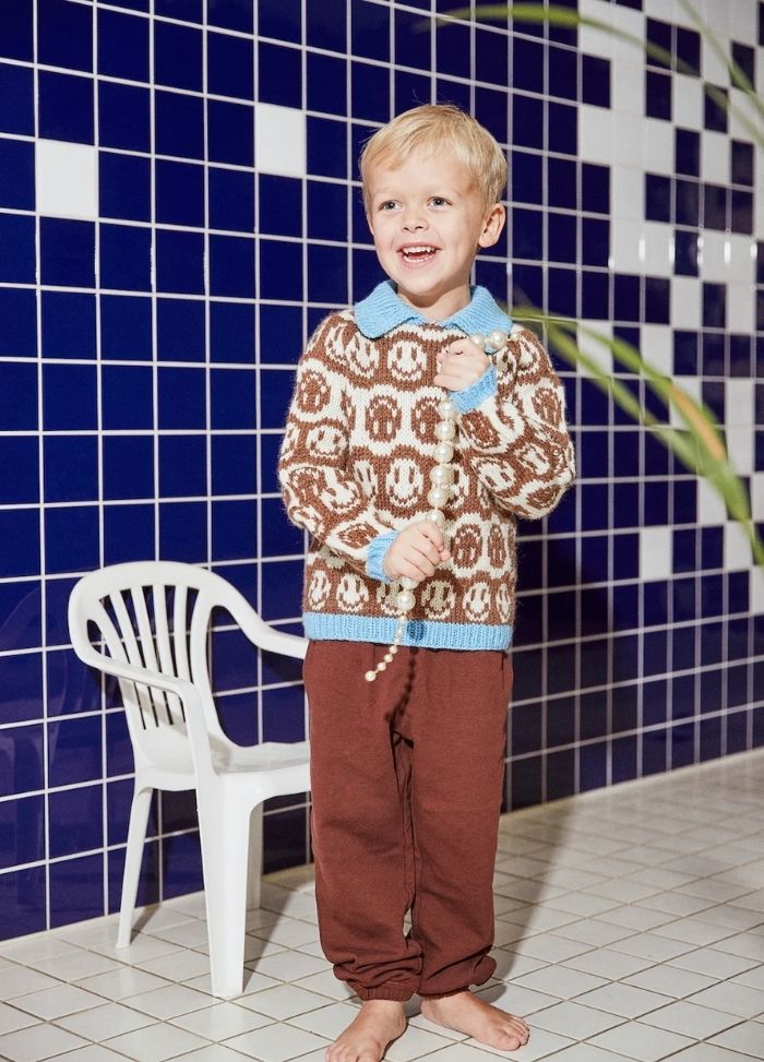 A smiling child in a sweater with smiley faces and red pants holds a beaded string, standing on a tiled floor with a blue and white tiled wall and a white plastic chair.