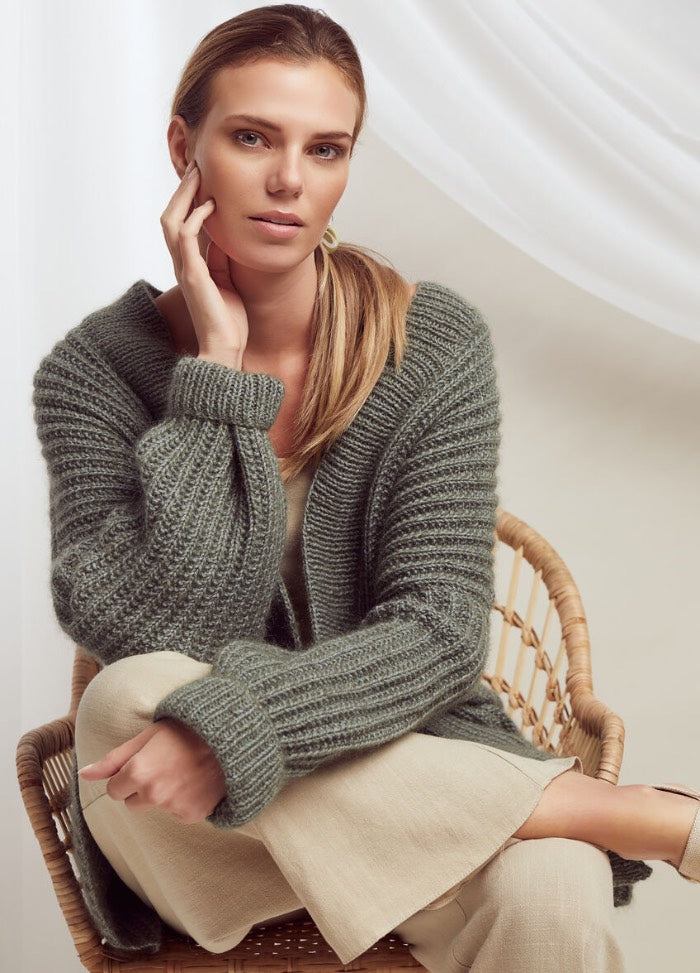A woman with a relaxed expression is seated in a wicker chair, wearing a chunky knit sweater and light pants. The setting is minimalist and serene.