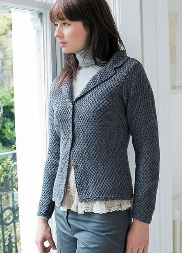 A woman with brown hair gazes out a window, wearing a textured gray jacket over a light blouse and turtleneck, conveying a serene, thoughtful mood.