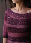 A woman wears a maroon, knitted sweater with intricate leaf patterns and shimmering sequins on the neckline. The mood is elegant and cozy.