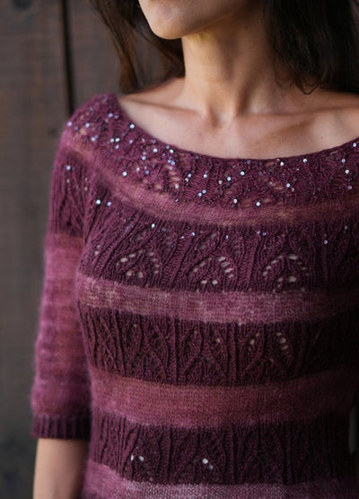 A woman wears a maroon, knitted sweater with intricate leaf patterns and shimmering sequins on the neckline. The mood is elegant and cozy.