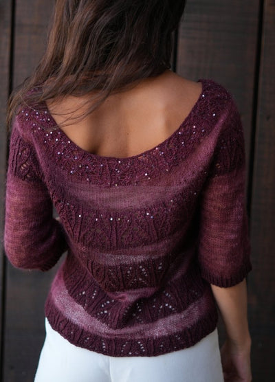 Woman facing away, wearing a maroon knit sweater with intricate patterns and small beads, paired with white pants. Her hair is loose, set against a dark wooden backdrop.