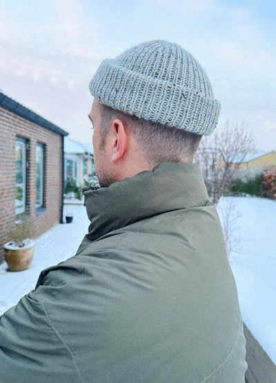 A person in an olive green jacket and grey knit cap stands outdoors on a snowy day, with a brick building and leafless trees in the background.