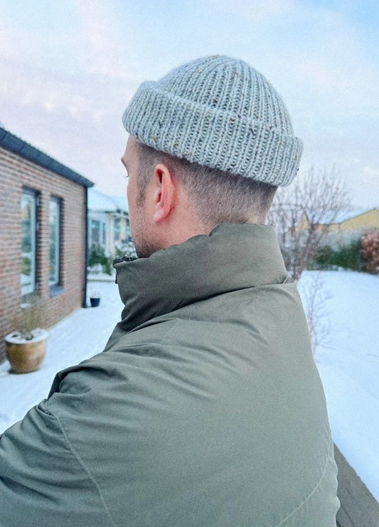 A person in an olive green jacket and grey knit cap stands outdoors on a snowy day, with a brick building and leafless trees in the background.