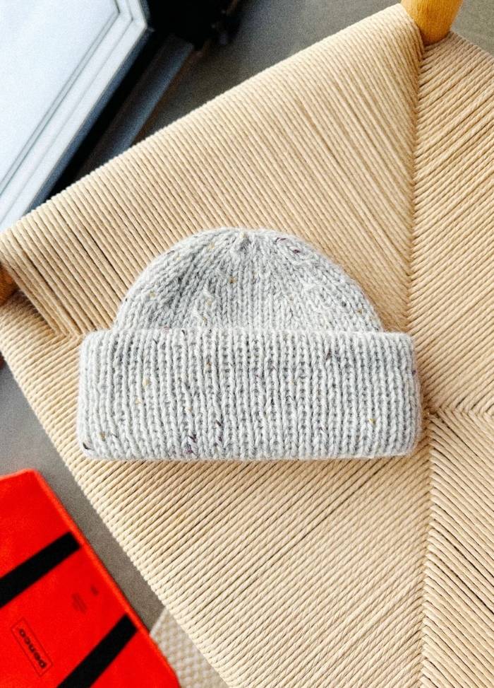 A grey knitted beanie lies atop a woven chair with a geometric pattern. The scene conveys a cozy, minimalist vibe with a red book in the corner.