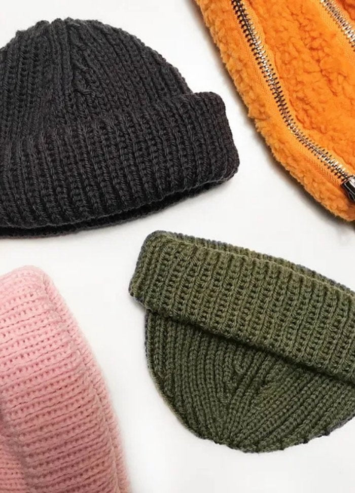 A collection of knitted hats and a jacket. The black and green hats are placed near a pink one, alongside an orange sherpa jacket with a zipper.