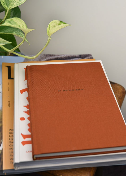 An orange notebook titled "MY KNITTING NOTES" lies on a stack of books on a wooden table. A green potted plant sits beside them, adding a cozy touch.