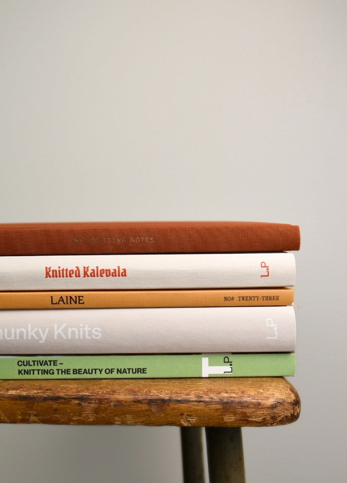 A stack of five books with neutral-toned covers sits on a rustic wooden stool against a plain background. The scene conveys a calm, cozy atmosphere.