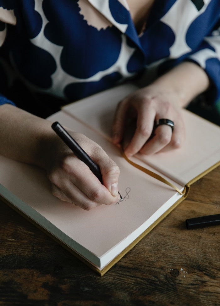 Person writing with a black pen in an open notebook on a wooden table. They wear a black ring and a blue patterned blouse, conveying focus.