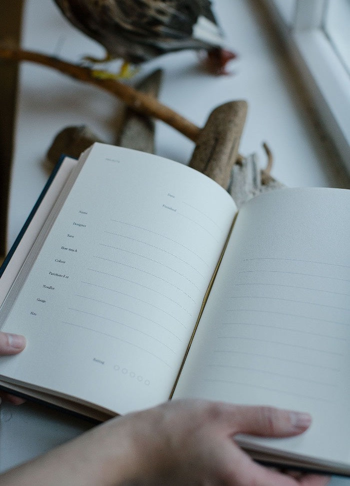 Open book with blank lined pages held by hands, resting near a window. A bird sculpture and driftwood are visible in the background, creating a calm ambiance.
