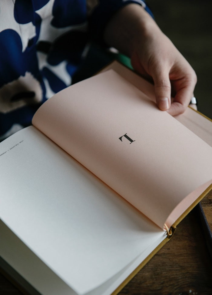 A hand is turning a pink book page with the letter "L." The person wears a blue and white patterned shirt. The scene is calm and focused.