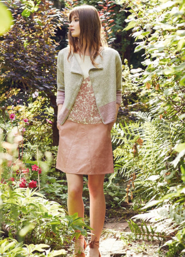 Model wearing the Nambu Jacket from the Rowan Softyak DK Collection by Marie Wallin, a light-colored knit jacket with pink and green tones layered over a floral top and paired with a light pink skirt, photographed in a vibrant garden filled with plants and flowers.