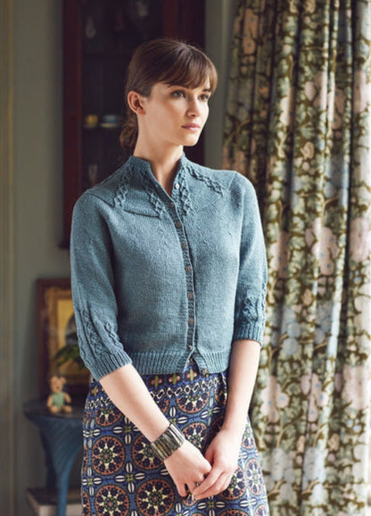 Model wearing the Norbu Cardigan from the Rowan Softyak DK Collection by Marie Wallin, a blue button-down knit with intricate stitch patterns on the shoulders and sleeves, paired with a geometric patterned skirt and a large bracelet, photographed indoors with floral curtains and decorative furniture.