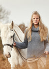 Person wearing the New Forest Sweater, a grey knit design, standing beside a white horse in a wooded outdoor setting.