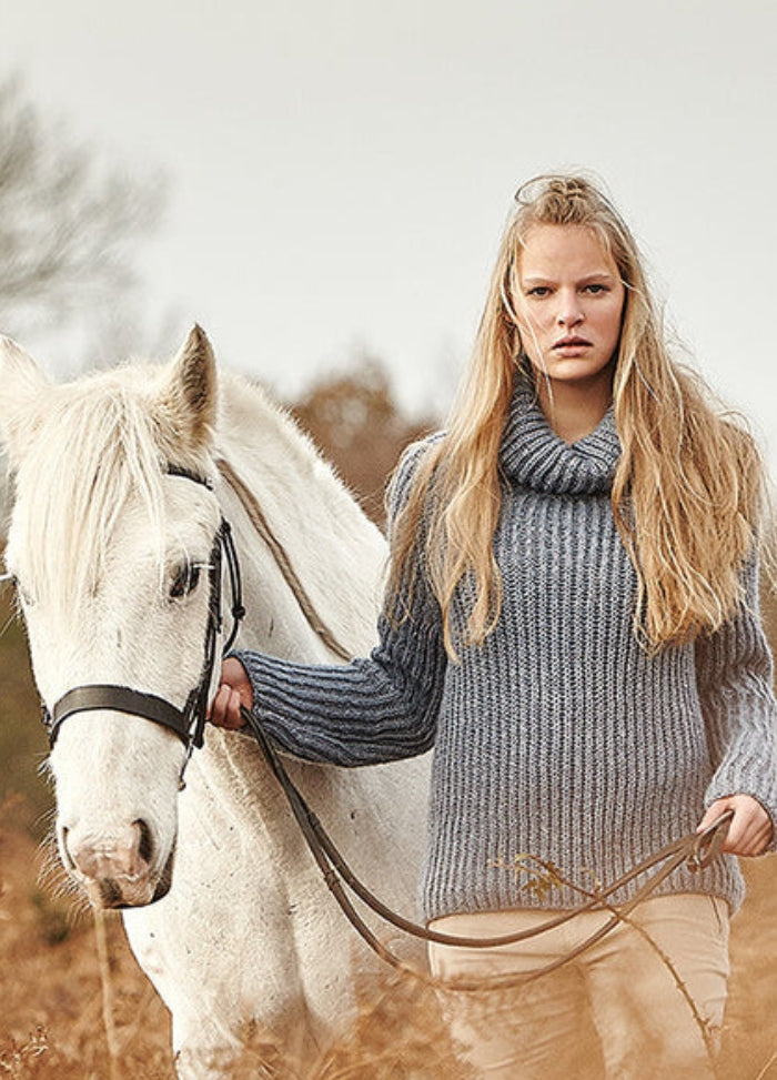 Person wearing the New Forest Sweater, a grey knit design, standing beside a white horse in a wooded outdoor setting.