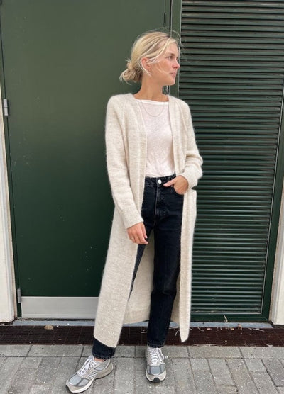 Woman in a cozy, cream cardigan over a white top and black jeans stands in front of a dark green door. Casual, relaxed pose, and calm atmosphere.