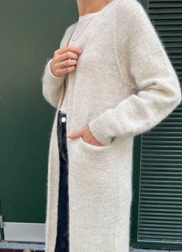 A person wears a cozy, long beige cardigan over a white top and dark jeans. One hand is in a pocket, conveying a relaxed and comfortable vibe.