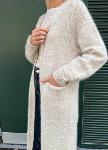 A person wears a cozy, long beige cardigan over a white top and dark jeans. One hand is in a pocket, conveying a relaxed and comfortable vibe.