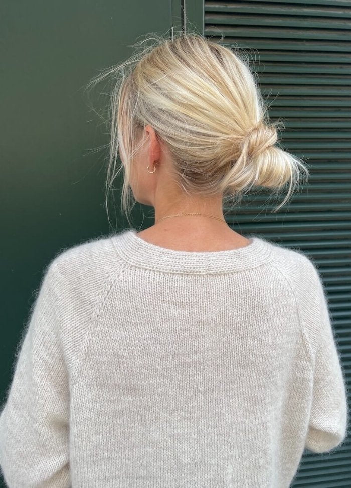 A person with blonde hair in a loose bun wears a light beige knit sweater, standing against a green wall with horizontal slats, exuding a relaxed, cozy vibe.