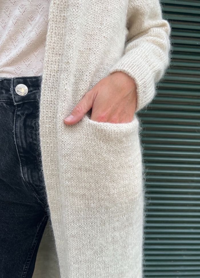 A hand partially in the pocket of a cozy, cream-coloured knit cardigan. The person wears a textured white top and dark jeans, exuding warmth and comfort.